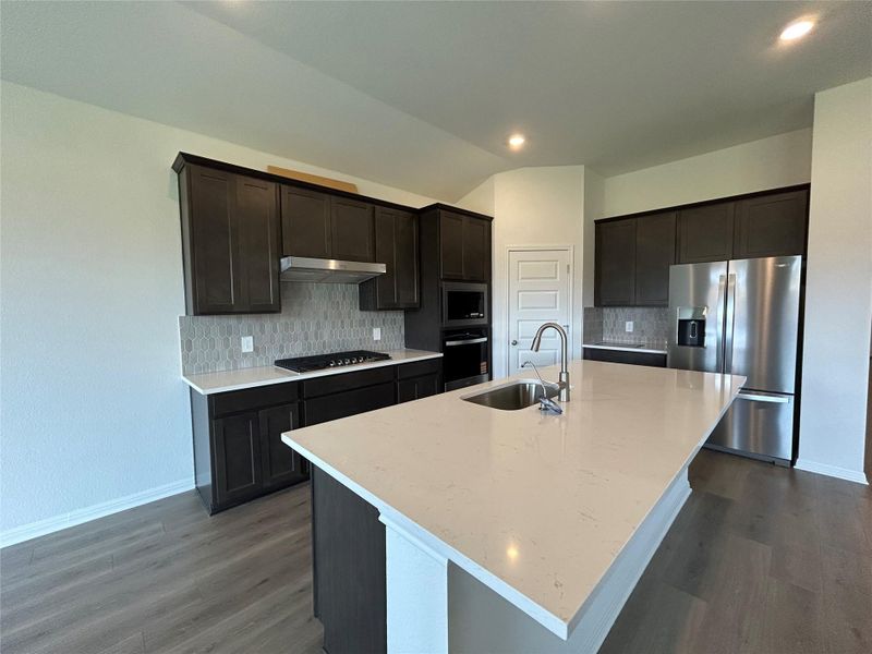 Kitchen featuring tasteful backsplash, appliances with stainless steel finishes, light stone countertops, an island with sink, and dark wood finished floors Kitchen featuring tasteful backsplash, appliances with stainless steel finishes, light stone countertops, an island with sink, and dark wood finished floors