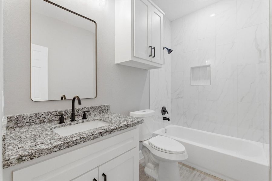 Bright and modern secondary bathroom featuring a soaking tub with floor-to-ceiling marble-patterned tile and a built-in shampoo niche. The space is elevated by a granite-topped vanity with an undermount sink, complemented by sleek matte black fixtures and a contemporary framed mirror.