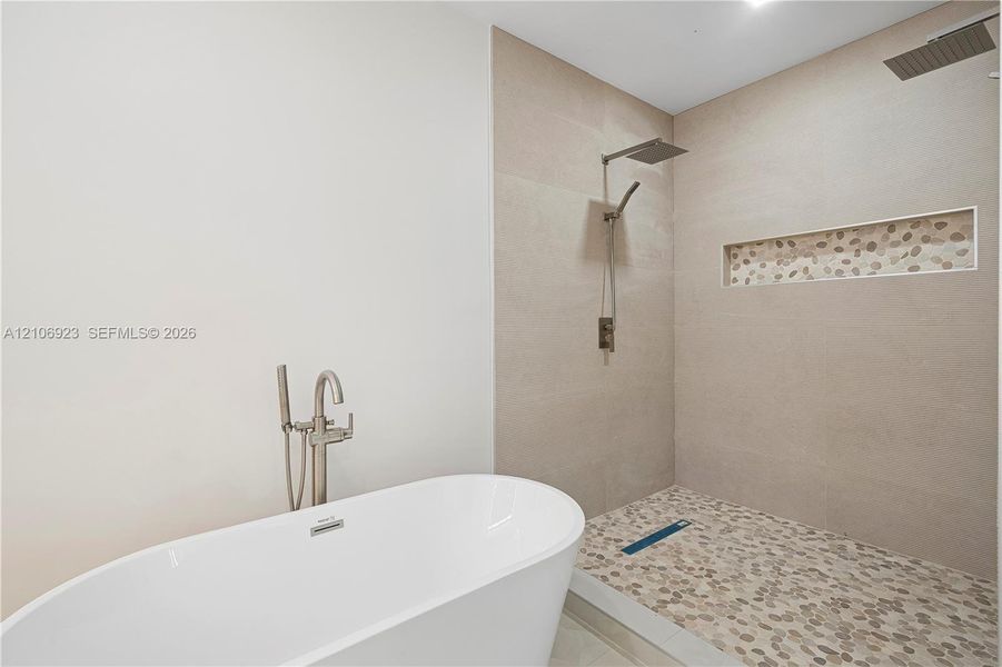 Freestanding soaking tub with a floor-mounted brushed nickel faucet, walk-in shower featuring large-format wall tile, pebble-stone flooring, rainfall showerhead, and a built-in tiled shower niche Freestanding soaking tub with a floor-mounted brushed nickel faucet, walk-in shower featuring large-format wall tile, pebble-stone flooring, rainfall showerhead, and a built-in tiled shower niche