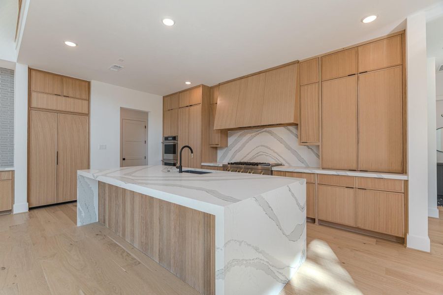 This modern kitchen features sleek wooden cabinetry, a large marble island with a built-in sink, and high-end appliances. The light wood floors and recessed lighting enhance the open, airy feel.