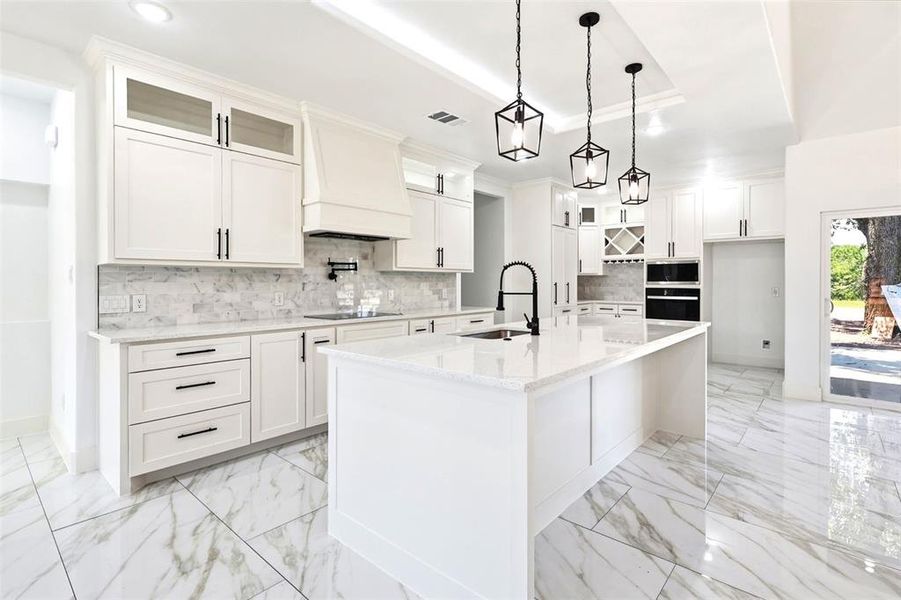 Modern kitchen featuring a large central island with a sink, white cabinetry with black hardware, marble-look tile flooring, and a stone backsplash