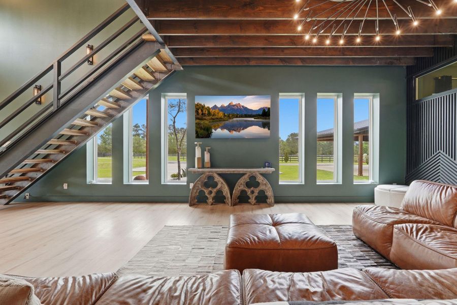 This spacious living room features high ceilings with exposed beams, a modern staircase, and large windows offering plenty of natural light and views of the outdoors. The open design includes a stylish chandelier and a cozy seating area with leather couches, creating a warm and inviting atmosphere.