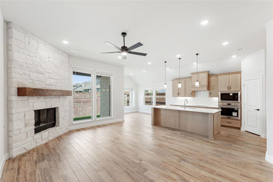 Open living area with stone fireplace flowing into kitchen with island and light wood cabinets