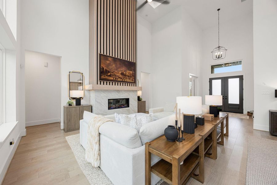 This spacious living room features high ceilings and abundant natural light. It includes a modern fireplace with a wood accent wall, elegant lighting, and stylish neutral decor. The open layout seamlessly connects to the entryway, creating an inviting atmosphere.