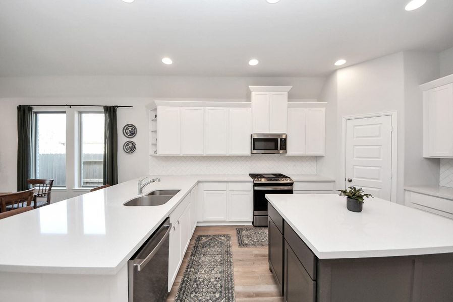 This beautifully designed kitchen features crisp white cabinetry, sleek stainless steel appliances, and a spacious island with an integrated sink and elegant countertops. Large windows fill the space with natural light, while recessed lighting enhances the bright, modern atmosphere.