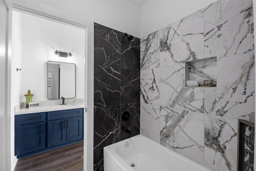 Stylish mix of black and white marble tiles around the bathtub, with a convenient built-in shelf. The adjacent vanity area includes a modern mirror, bright lighting, and a striking blue cabinet, complemented by a sleek countertop.
