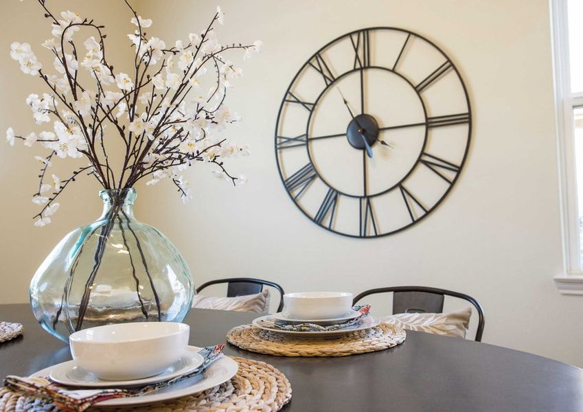 Wooden table with placemats, white bowls and glass ornament with flowers plus large wall clock in the back.