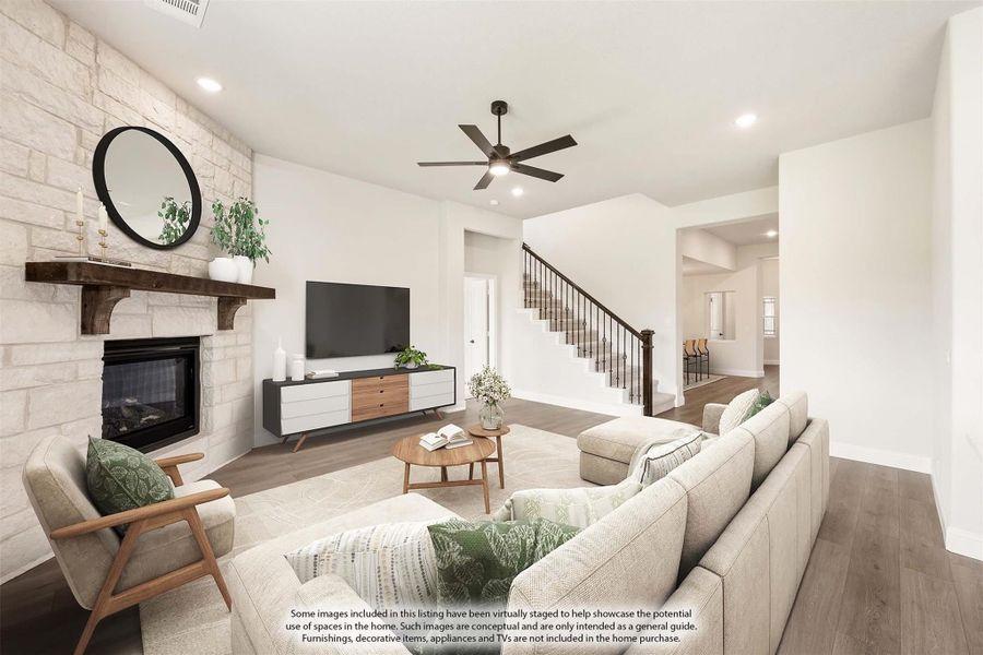 Living room with stone fireplace, ceiling fan, hardwood floors, and staircase in background