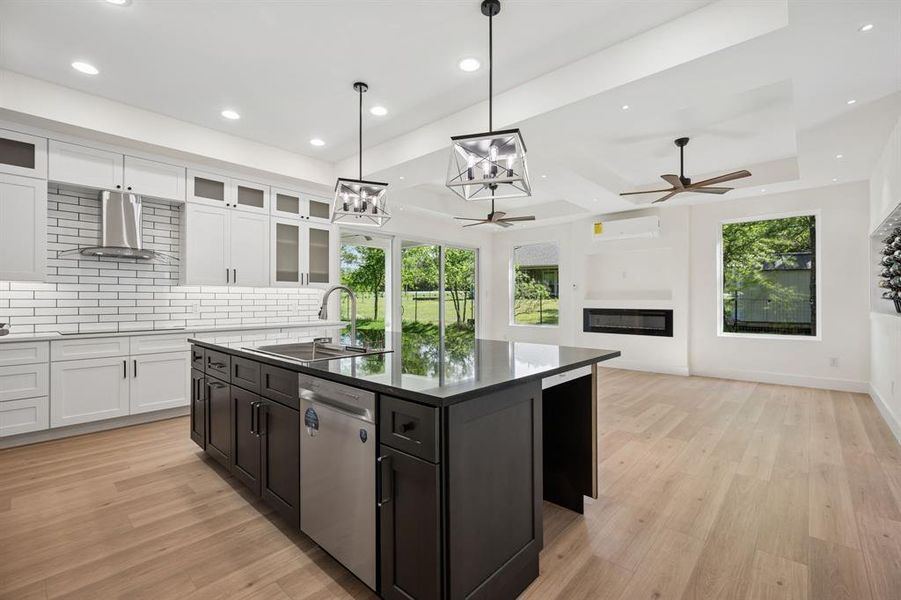 Two tone kitchen with a kitchen island with sink, a glass covered fireplace, light wood finished floors, stainless steel dishwasher, and two tone cabinets. Space for two undercabinet refrigerators.