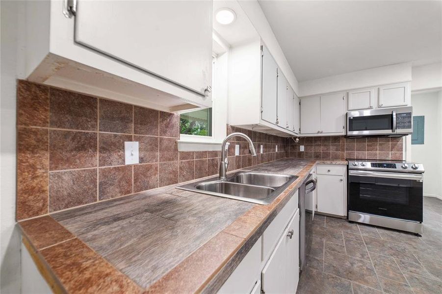Kitchen with tile countertops, backsplash, appliances with stainless steel finishes, white cabinets, and a sink Kitchen with tile countertops, backsplash, appliances with stainless steel finishes, white cabinets, and a sink