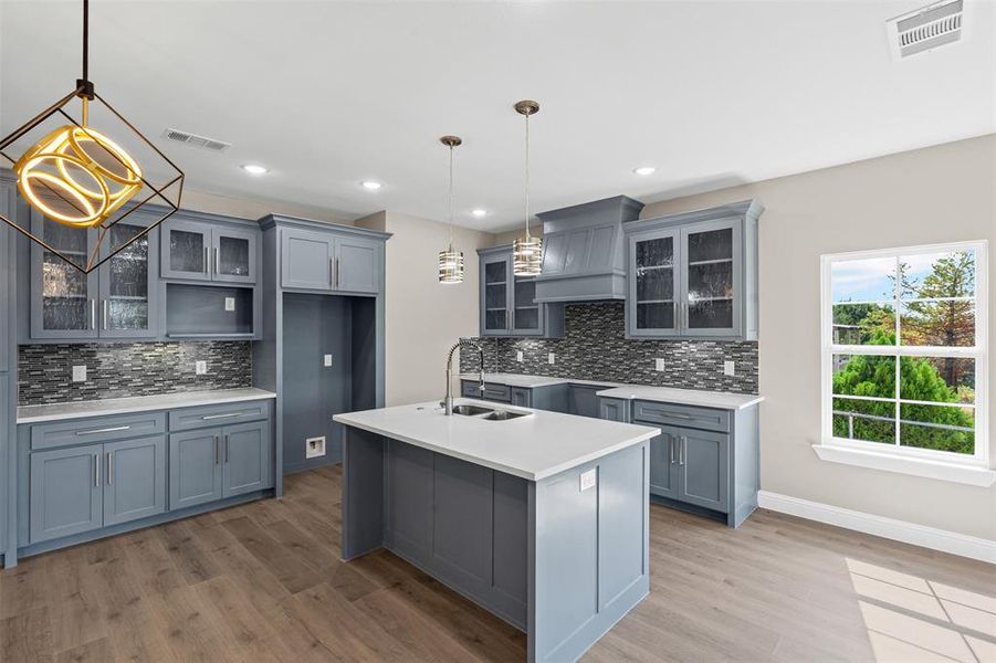 Kitchen with glass insert cabinets, light wood finished floors, custom range hood, and recessed lighting Kitchen with glass insert cabinets, light wood finished floors, custom range hood, and recessed lighting