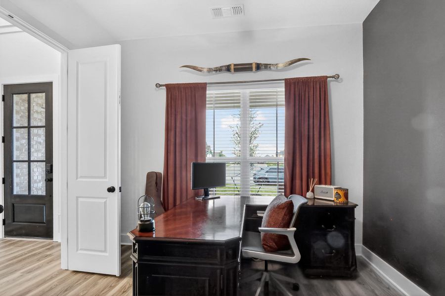Room featuring wood-finish flooring, a window with horizontal blinds and drapery, a solid panel interior door, and a multi-pane glass-insert exterior door Room featuring wood-finish flooring, a window with horizontal blinds and drapery, a solid panel interior door, and a multi-pane glass-insert exterior door