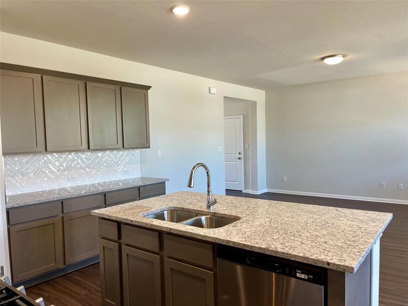 Kitchen featuring dishwasher, light stone counters, dark wood-style floors, decorative backsplash, and an island with sink