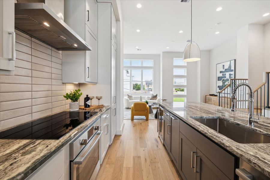 This modern kitchen features sleek cabinetry, a polished marble countertop, and a built-in cooktop. It opens into a bright living area with large windows, providing abundant natural light and a spacious, airy feel.