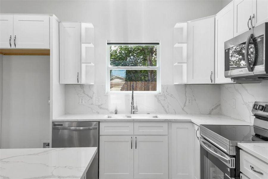 Modern kitchen featuring white shaker cabinetry, stainless steel appliances, a white stone countertop, a matching stone backsplash, and a large window above the sink