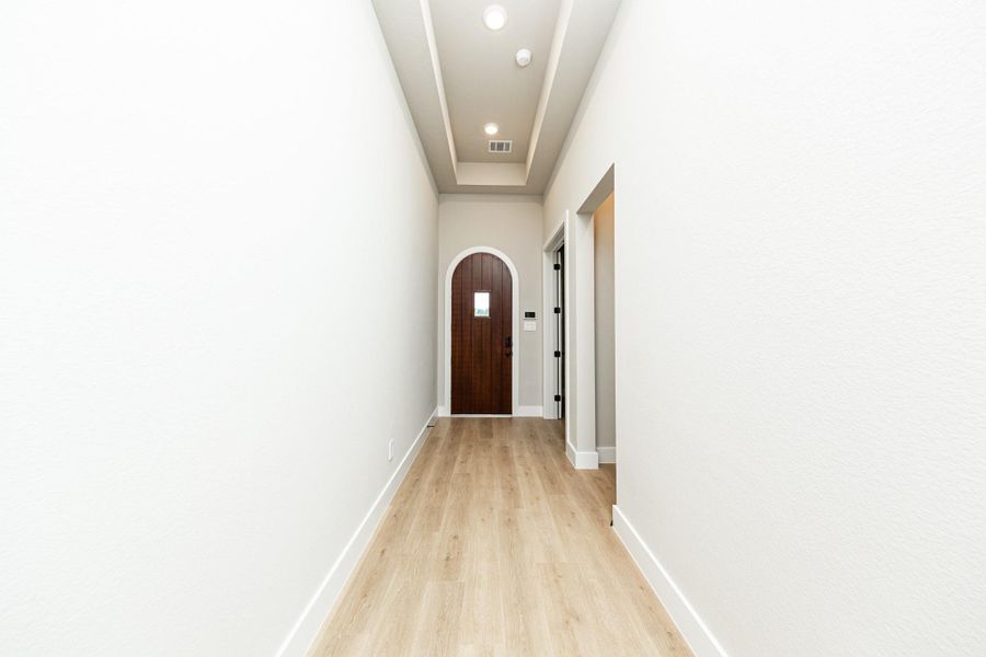 This long entryway features wood-look flooring and a tray ceiling. This Spanish-style front door serves as a bold architectural focal point, featuring a classic round arch top and a rich, dark-stained wood finish that provides a deep contrast against the home's light-colored walls This long entryway features wood-look flooring and a tray ceiling. This Spanish-style front door serves as a bold architectural focal point, featuring a classic round arch top and a rich, dark-stained wood finish that provides a deep contrast against the home's light-colored walls