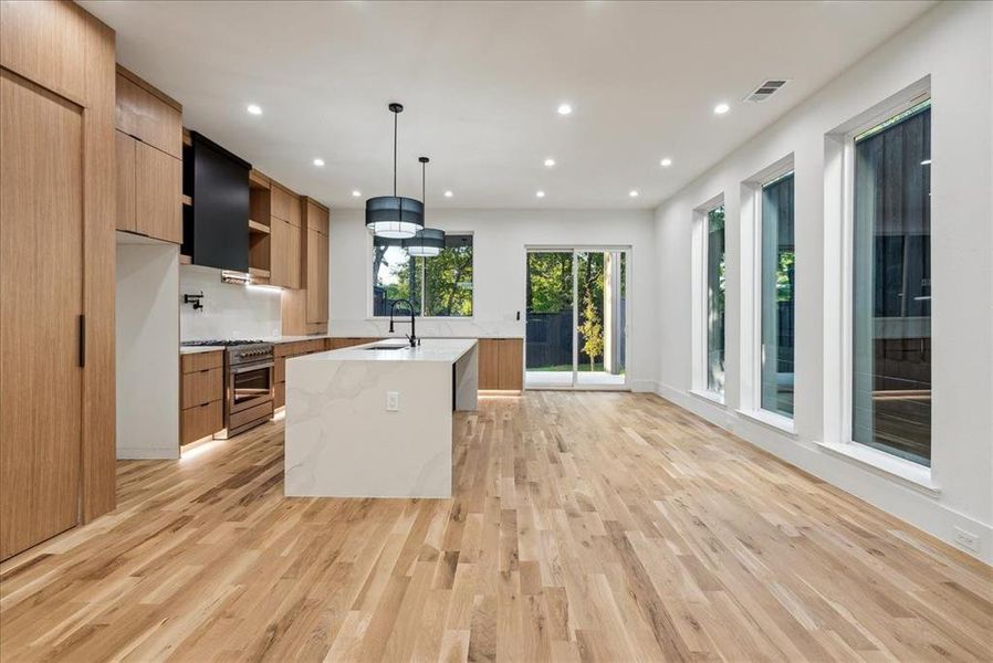 Kitchen with light hardwood / wood-style floors, stainless steel stove, a center island with sink, decorative light fixtures, and light stone counters