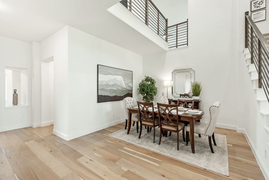 Formal dining room with two-story ceiling, wood dining table, six chairs, and light hardwood floors near staircase railing.
