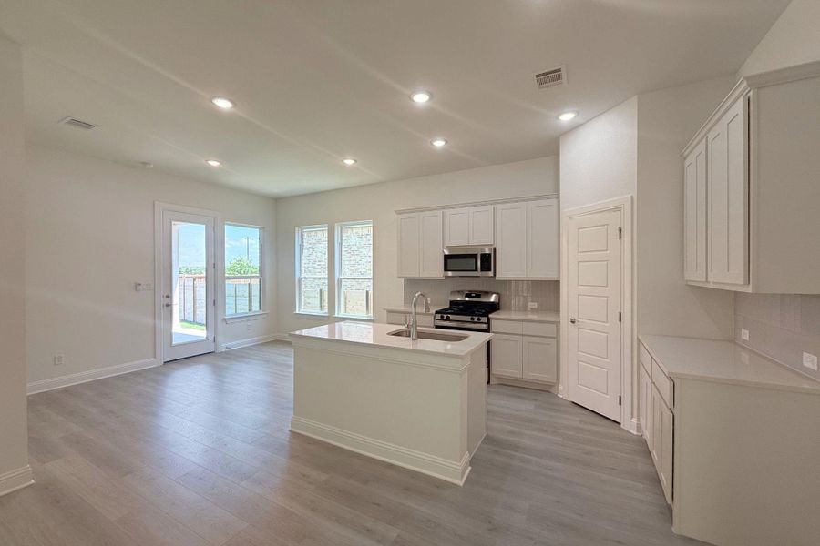 Open-concept kitchen featuring white shaker cabinetry, stainless steel appliances, a center island with sink, and light wood-finish flooring
