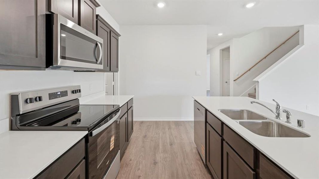 Kitchen with stainless steel appliances, light wood finished floors, dark brown cabinetry, recessed lighting, and light stone counters