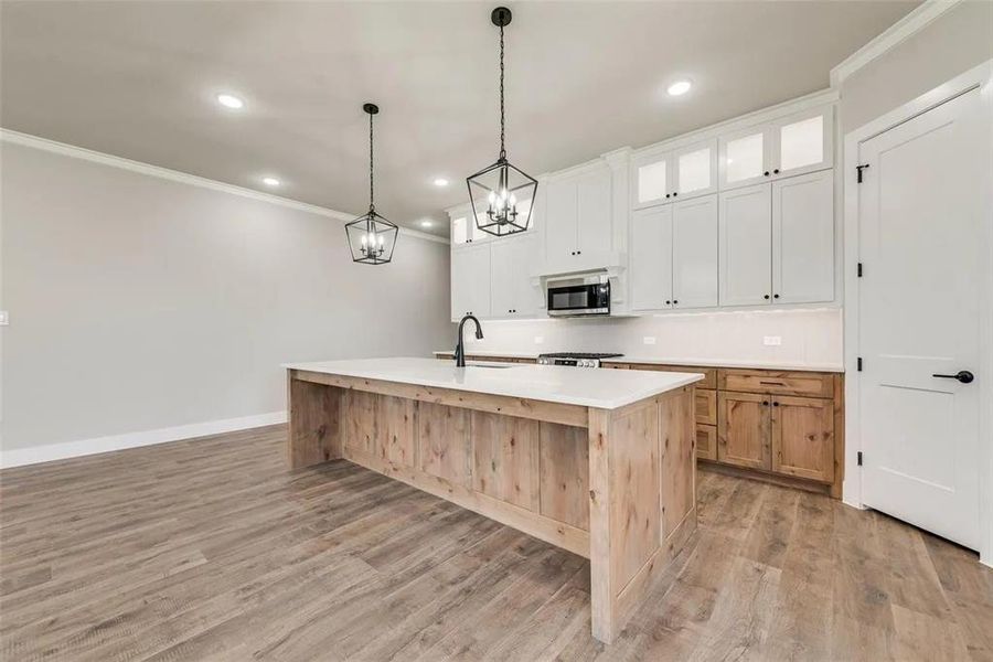 Kitchen featuring two tone cabinets, ornamental molding, an island with sink, glass insert cabinets, and light wood-style flooring
