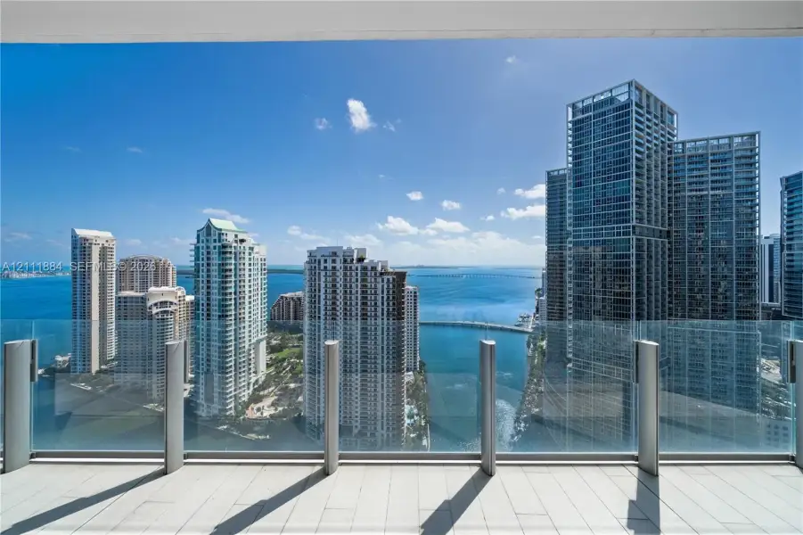 Covered balcony featuring tile flooring and a glass panel railing, showcasing an expansive waterfront and city skyline panorama Covered balcony featuring tile flooring and a glass panel railing, showcasing an expansive waterfront and city skyline panorama