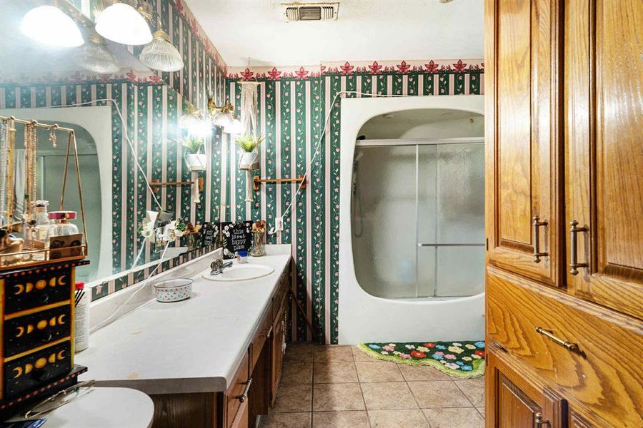 Bathroom featuring vanity, light tile patterned flooring, wallpapered walls, and shower / bath combination with glass door Bathroom featuring vanity, light tile patterned flooring, wallpapered walls, and shower / bath combination with glass door