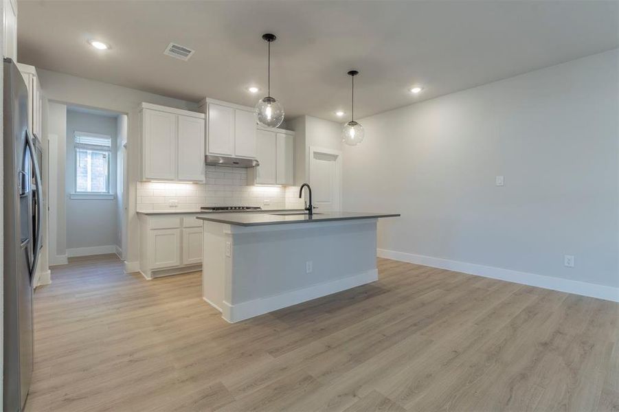 Kitchen with an island with sink, dark countertops, stainless steel appliances, decorative light fixtures, and light wood-style flooring