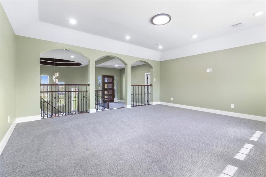 This photo showcases a spacious, bright room with soft green walls and modern lighting. It features an open layout with carpet flooring and decorative arches leading to a staircase with wrought iron railings, enhancing the home's elegance.