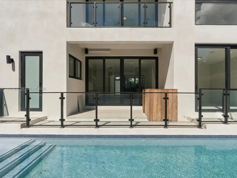 Exterior rear patio featuring smooth stucco finish, black-framed sliding glass doors, glass panel railings, and an integrated swimming pool with tiled steps Exterior rear patio featuring smooth stucco finish, black-framed sliding glass doors, glass panel railings, and an integrated swimming pool with tiled steps