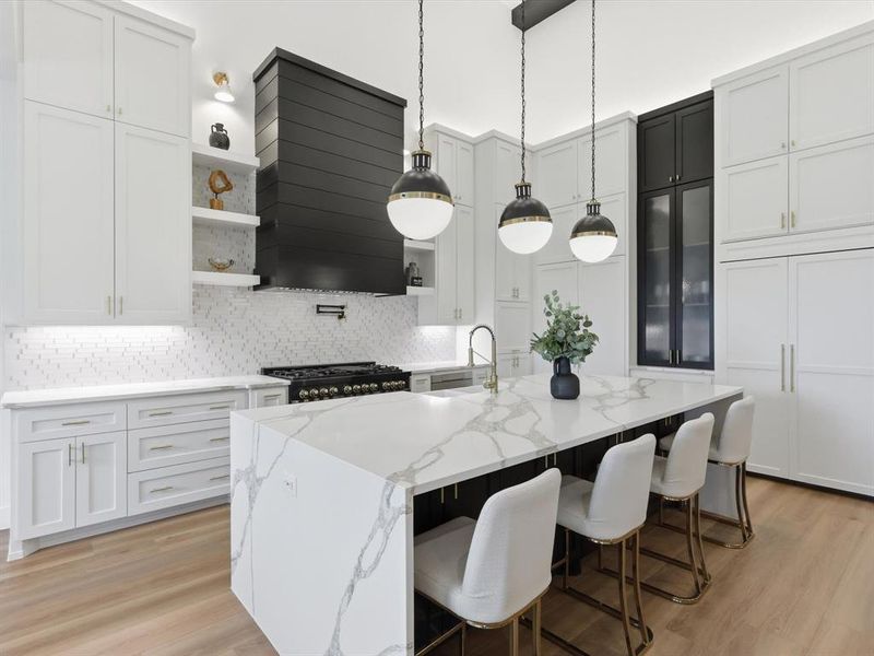Gourmet kitchen featuring a large central island with waterfall-edge countertop, extensive white cabinetry, a black range hood, a pot filler, and light wood-finish flooring