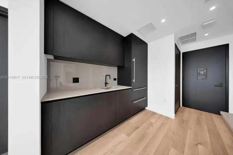Modern kitchenette featuring matte black flat-panel cabinetry, integrated refrigerator, neutral solid-surface countertop, light wood-finish flooring, and a matte black faucet Modern kitchenette featuring matte black flat-panel cabinetry, integrated refrigerator, neutral solid-surface countertop, light wood-finish flooring, and a matte black faucet