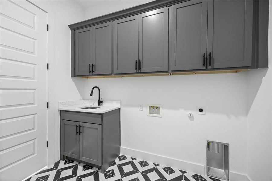 Laundry room featuring a white paneled door, dark gray shaker-style cabinetry, a white countertop with integrated sink, a matte black faucet, and patterned floor tiling