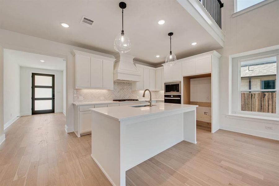 Kitchen featuring backsplash, a center island with sink, hanging light fixtures, white cabinets, and light wood-style flooring Kitchen featuring backsplash, a center island with sink, hanging light fixtures, white cabinets, and light wood-style flooring