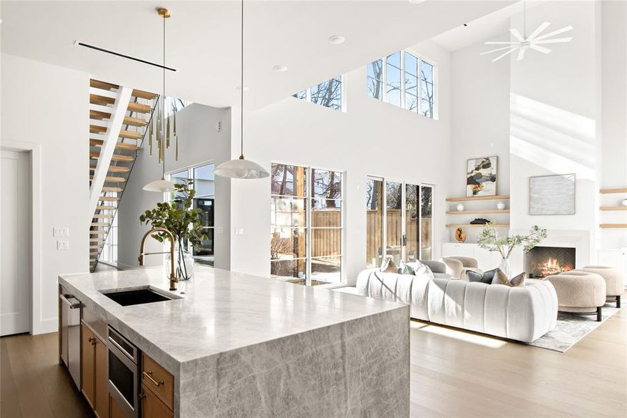 Open-concept living space featuring a waterfall island with an undermount sink, a two-story ceiling, multiple large windows, and a modern fireplace