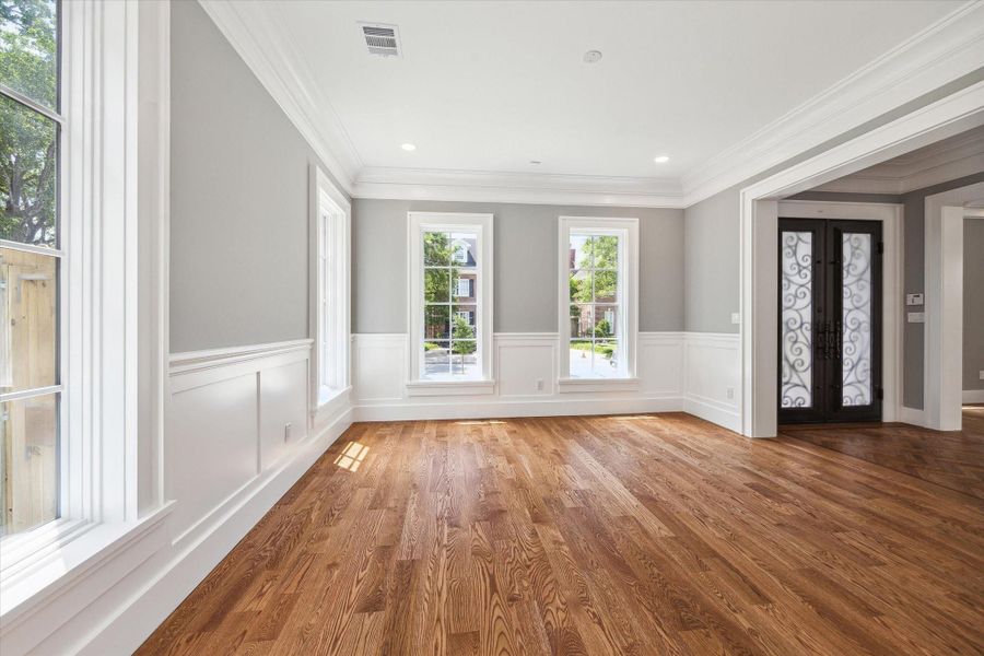 This photo shows the bright and spacious dining room with hardwood floors, large windows, and elegant crown molding. The neutral gray walls and white wainscoting add a touch of sophistication, and the decorative front door enhances the entryway as you look from the dining room.