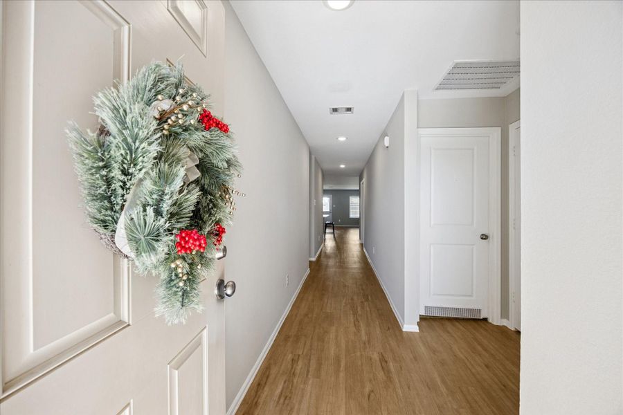 This photo shows a welcoming entry hallway with light flooring and neutral walls. A decorative wreath adorns the front door, adding a festive touch. The hallway leads to a bright living area at the far end, suggesting a spacious and inviting home layout.