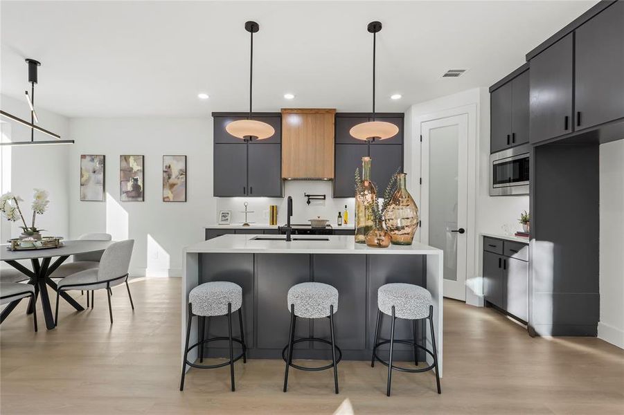 Kitchen with hanging light fixtures, an island with sink, a breakfast bar, light wood-type flooring, and light stone counters
