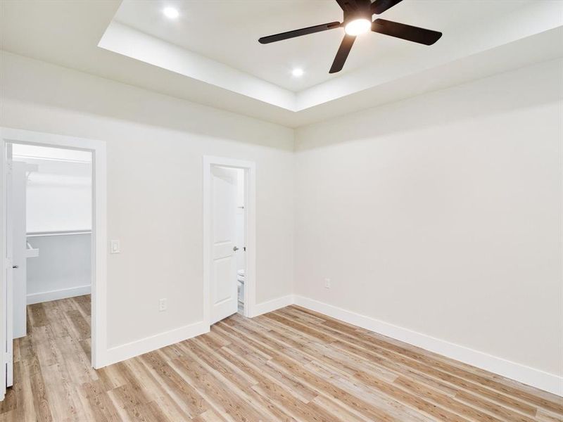 Spacious room featuring wood-finish flooring, a tray ceiling with recessed lighting, and a dark-toned ceiling fan Spacious room featuring wood-finish flooring, a tray ceiling with recessed lighting, and a dark-toned ceiling fan