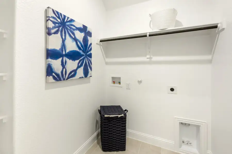 Laundry room with white walls, upper shelf with hanging rod, washer hookups, and a dark wicker hamper on tile floor