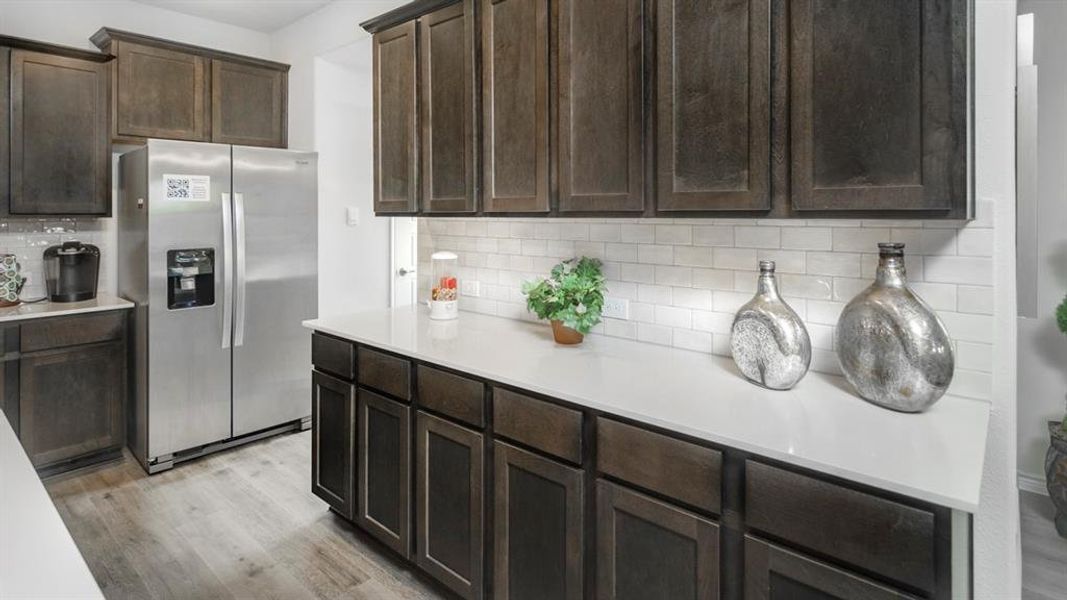 Kitchen featuring dark wood-finish cabinetry, light-toned countertops, subway tile backsplash, stainless steel refrigerator, and wood-finish flooring