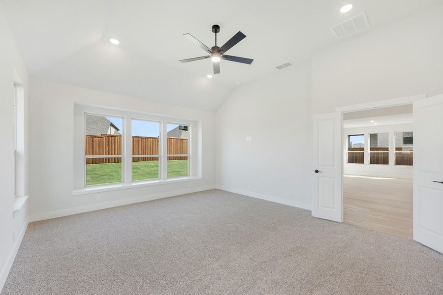 Empty bedroom with vaulted ceiling, ceiling fan, carpet flooring, and large windows overlooking backyard