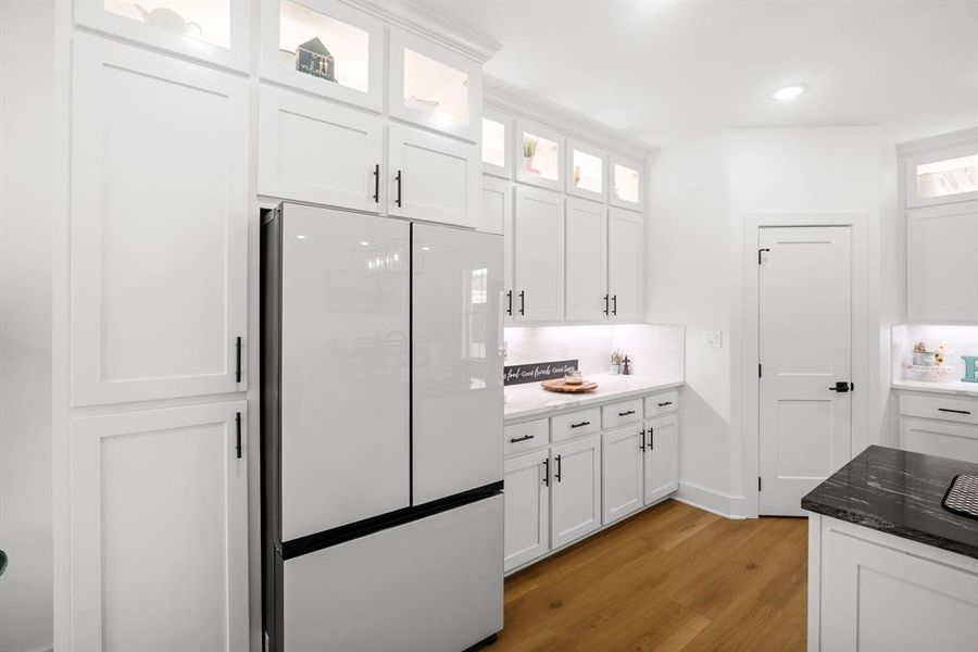 Kitchen with freestanding refrigerator, light wood-style flooring, white cabinets, dark stone countertops, and glass insert cabinets Kitchen with freestanding refrigerator, light wood-style flooring, white cabinets, dark stone countertops, and glass insert cabinets