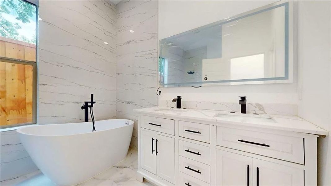 Freestanding soaking tub with matte black floor-mounted faucet, dual vanity with undermount sinks, quartz countertops, and an illuminated mirror