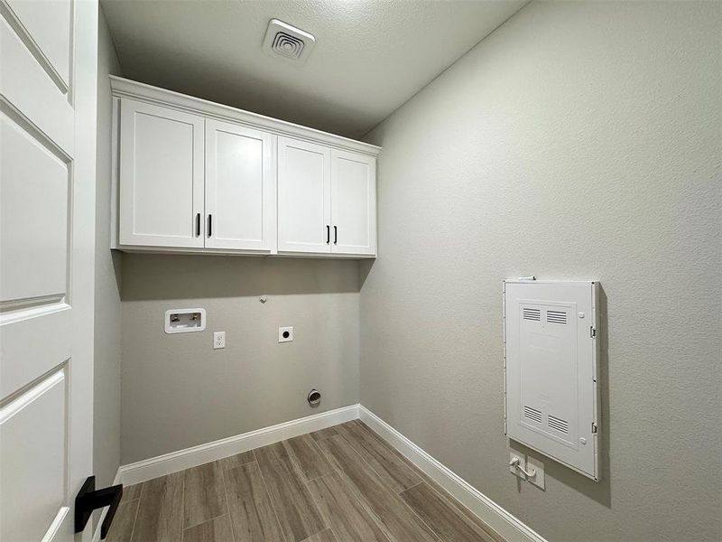 Dedicated laundry space featuring wood-finish flooring, white shaker-style cabinetry with matte black hardware, and an overhead ventilation fan