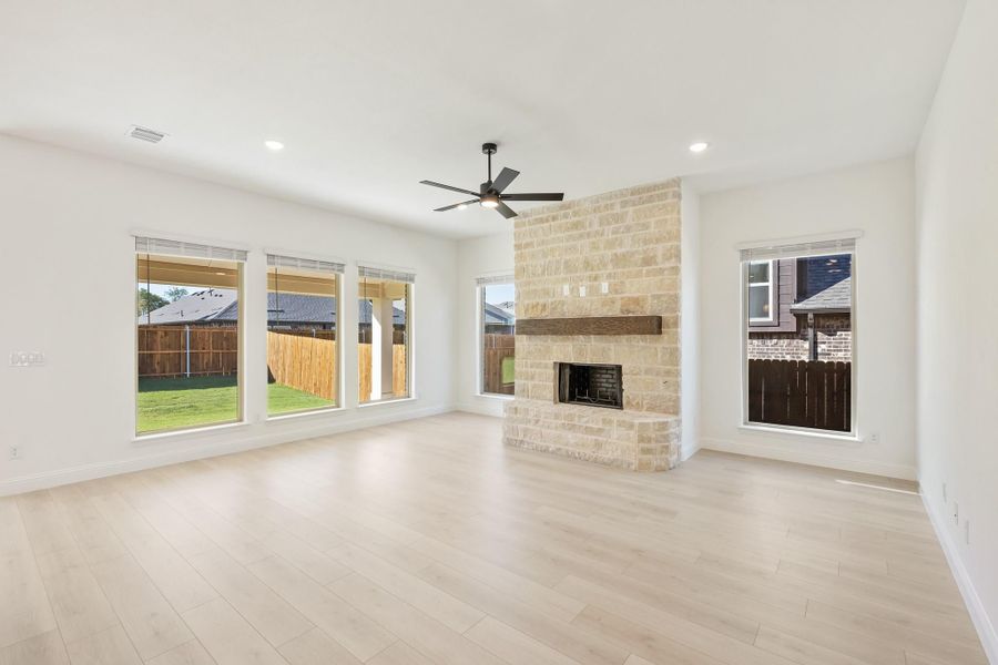 Open living room with stone fireplace, light hardwood floors, ceiling fan, and large windows overlooking backyard Open living room with stone fireplace, light hardwood floors, ceiling fan, and large windows overlooking backyard