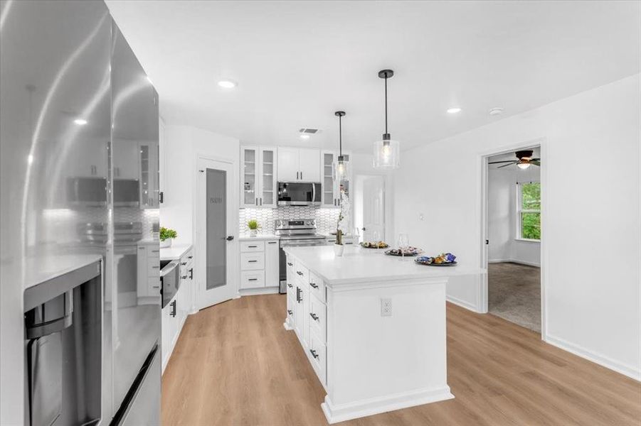 Kitchen featuring appliances with stainless steel finishes, light wood flooring, ceiling fan, white cabinetry, and a center island with sink