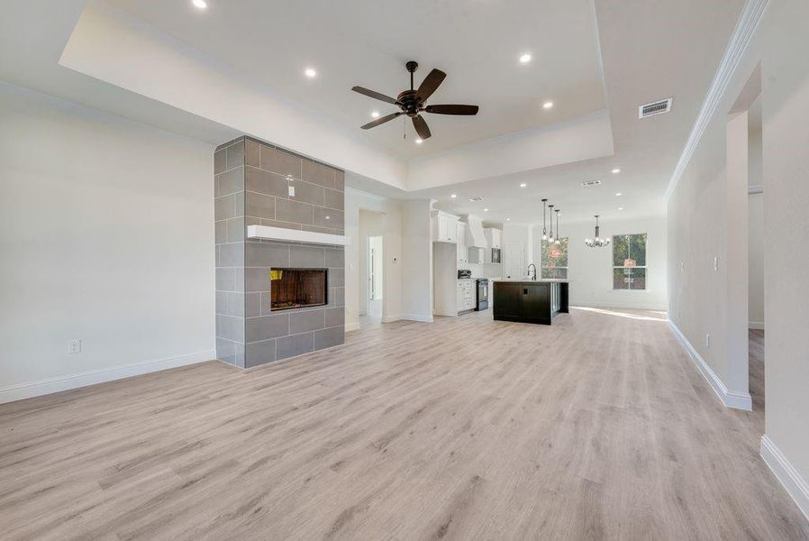 Unfurnished living room with ceiling fan with notable chandelier, a tiled fireplace, a tray ceiling, and light wood-type flooring