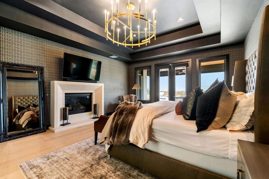Bedroom with wallpapered walls, a tray ceiling, a chandelier, wood finished floors, and a glass covered fireplace Bedroom with wallpapered walls, a tray ceiling, a chandelier, wood finished floors, and a glass covered fireplace