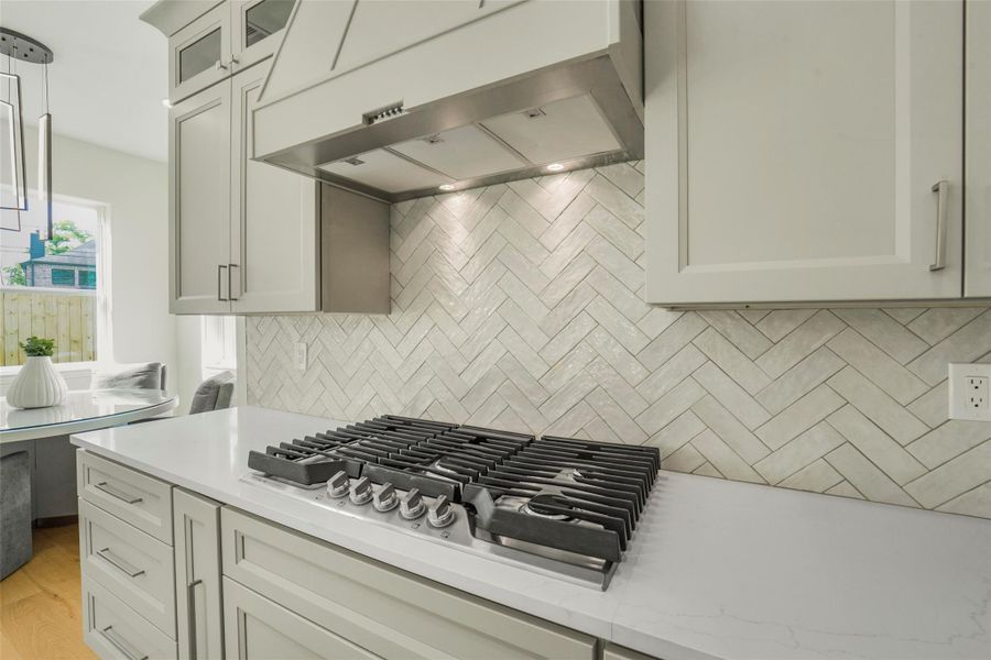 This kitchen features a sleek gas cooktop with a modern hood, light-colored cabinetry, and a stylish herringbone tile backsplash. Ample natural light and a nearby dining area create a bright, inviting space.